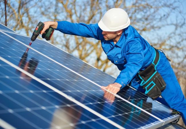 Auf dem Bild ist ein Arbeiter in einem blauen Overall zu sehen, der eine Photovoltaikanlage installiert.