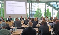Hier ist ein Foto von einem Konferenzraum zu sehen. Im Vordergrund sitzen mehrere Personen an einem langen rechteckigen Tisch mit dem Rücken zur Kamera. An der Stirnseite des Tisches sitzen weitere Personen, die zum Raum blicken. An der Wand hinter ihnen ist eine große Leinwand mit einer Präsentation zu sehen. Der Raum hat große Fenster, durch die man Gebäude und Bäume im Freien erkennen kann. Hier ist ein Foto von einem Konferenzraum zu sehen. Im Vordergrund sitzen mehrere Personen an einem langen rechteckigen Tisch mit dem Rücken zur Kamera. An der Stirnseite des Tisches sitzen weitere Personen, die zum Raum blicken. An der Wand hinter ihnen ist eine große Leinwand mit einer Präsentation zu sehen. Der Raum hat große Fenster, durch die man Gebäude und Bäume im Freien erkennen kann.