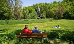 Ein Paar sitzt auf einer Bank und blickt auf die grüne Landschaft, in der Mitte ist ein Fels im Wald zu sehen. Ein Paar sitzt auf einer Bank und blickt auf die grüne Landschaft, in der Mitte ist ein Fels im Wald zu sehen.