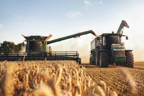 Hier ist ein Bild zu sehen: Im Vordergrund ist Weizen zu sehen und im Hintergrund zwei landwirtschaftliche Maschinen die den Weizen ernten. 